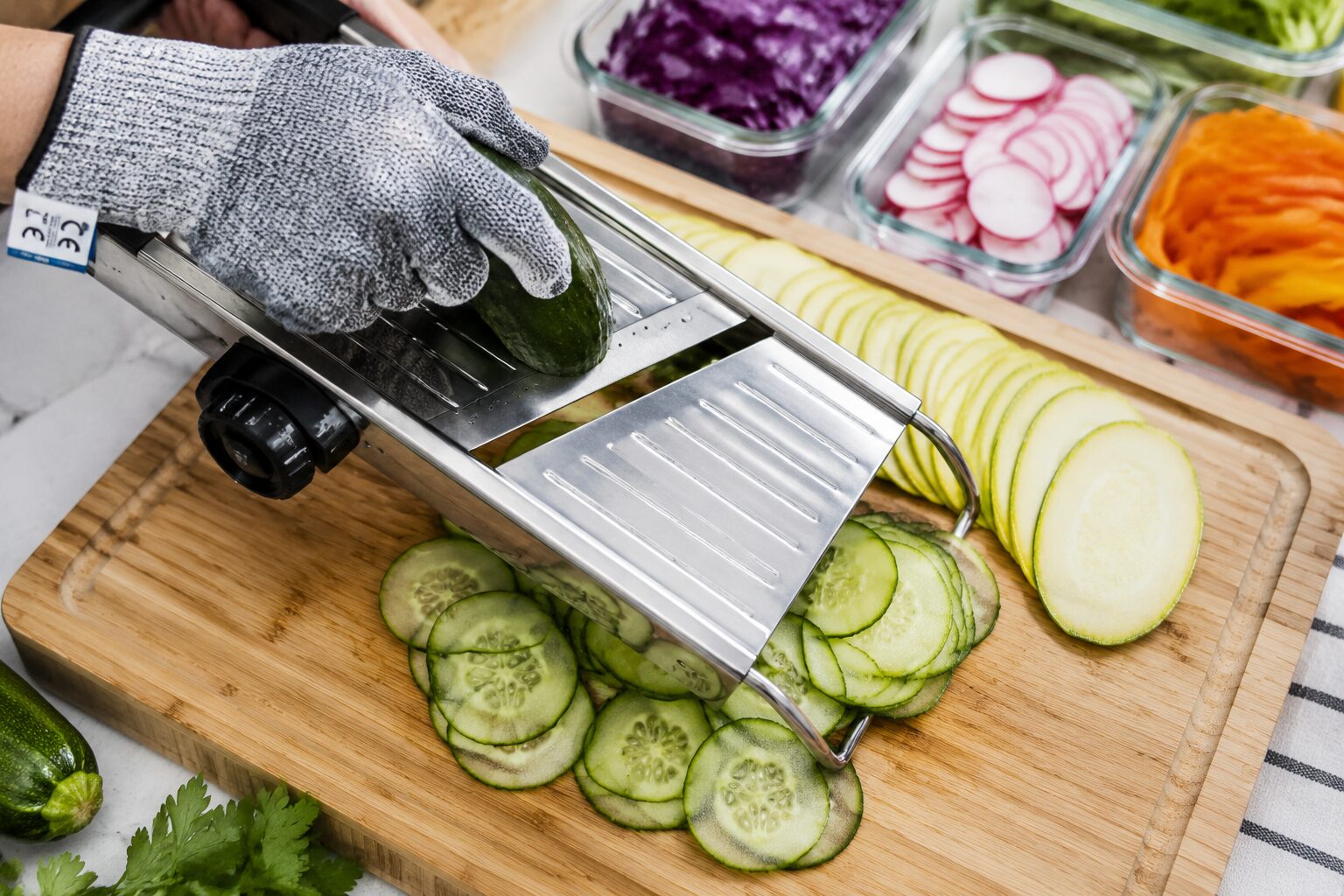 Close-up overhead shot of a stainless steel mandoline slicer in action on a wooden cutting board, with paper-thin cucumber