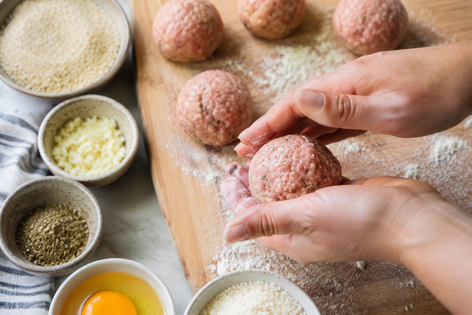 Close-up overhead shot of raw ground turkey meatballs being rolled by hand on a floured wooden cutting board, surrounded by